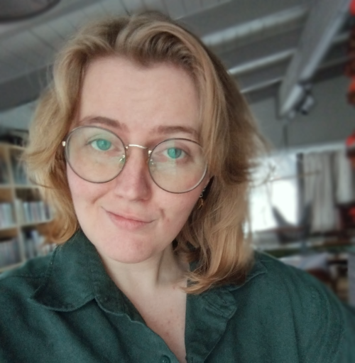 Photo of a nonbinary individual with round glasses and shoulder-length blond hair, wearing a teal button-down shirt and smirking warmly at the viewer.